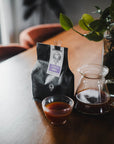 Coffee bag, glass of coffee, and coffee maker on a wooden table with a plant in the background.