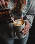 A person wearing a patterned sweater carefully pours steamed milk into a cup of coffee, creating intricate latte art. The scene feels cozy and artisanal.